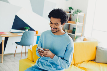 Young man casually dressed in blue sitting on couch at home, smiling and using his smartphone on a relaxing morning