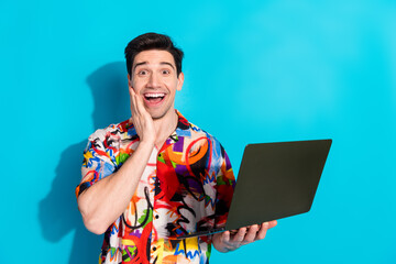 Cheerful young man wearing colorful shirt holding a laptop on a vibrant blue background showcasing...