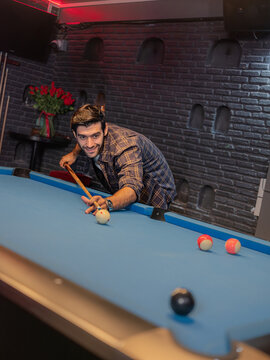 A focused young man lining up a pool shot in a modern bar. He leans over a blue-felt billiards table, hand steady on the cue stick, 