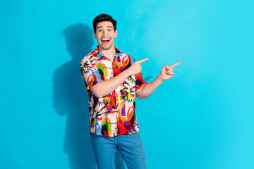 Cheerful young man in a multicolored shirt pointing enthusiastically against a bright blue...