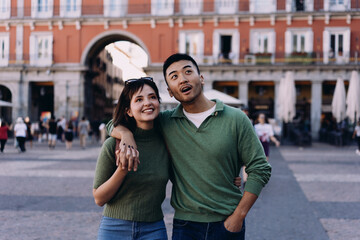 Smiling couple pausing in charming European square, looking up with awe and joy, embracing shared...
