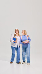 Fototapeta premium Full-length of two senior women with backpacks and books walking and talking giant white background. Elderly students. Concept of education for older adults, active aging