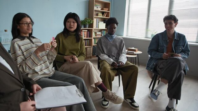 Slowmo pan shot of young biracial female school counselor holding paper clipboard speaking to group of multiethnic teenagers sitting on chairs in circle in cozy classroom listening to her