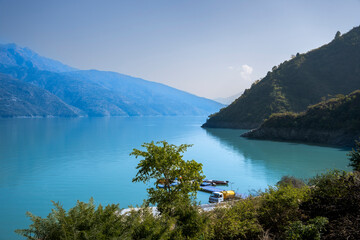 A truck is parked near the ferry jetty, with a lake and green-covered mountains surrounding it. This setting creates a functional yet scenic view.
