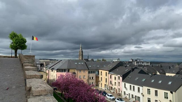 Panoramic, scenic skyline view of Arlon city buildings and Saint Martin church. Belgian flag is waving on top of Saint Donat church hill. Street with trees blooming in pink blossoms. Arlon, Belgium