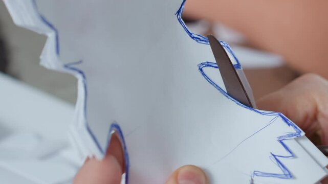 close hand view carefully using scissors cutting along blue outlined shapes on white paper while creating handmade craft project on table with scattered paper pieces during indoor creative session