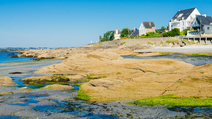 The coastline of Concarneau, Brittany, France, during low tide, with rocks and the waters of Atlantic ocean. Traditional residential houses on the right. Blue sky on the background.