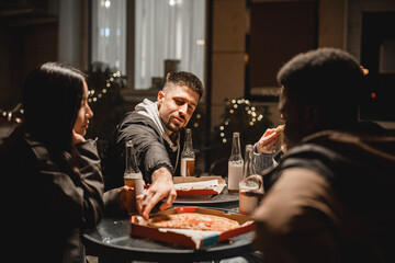 Friends sharing pizza and drinks at night in a cozy outdoor setting with festive lights