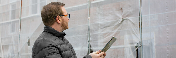 Caucasian male in glasses inspects construction site with digital tablet, urban development,...