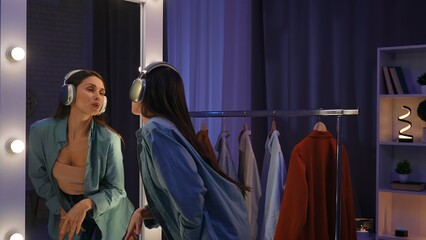 Older adult woman spending evening at home, standing in front of the mirror listening music in headphones, singing and dancing, positive expression.