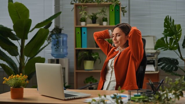 Older adult businesswoman works at office, woman sitting at desk working on laptop, sits back in chair with hands behind head, smiling positive expression.