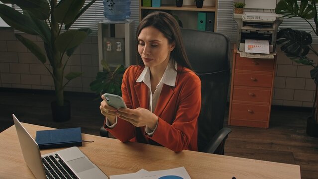 Older adult businesswoman works at green office with plants, woman sitting at desk working on laptop, scrolling social media on smartphone online. - Powered by Adobe