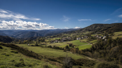 Fototapeta premium A picturesque countryside landscape reveals rolling hills with lush green fields and a small village led peacefully under a bright blue sky filled with soft clouds.