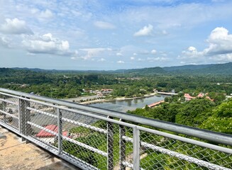 A scenic view from a lookout point featuring lush greenery, water bodies, and distant hills under a cloudy sky.