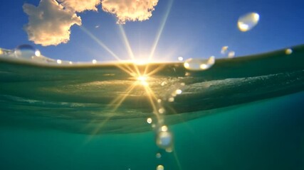 Underwater view of sunlight bursting through ocean waves. Turquoise water with sun rays and white clouds. Peaceful and serene atmosphere