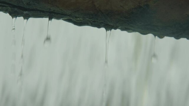 Close-up shot of water droplets falling from the edge of a stone lintel, with a soft-focus background of heavy rain cascading in sheets.