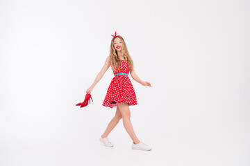 Joyful young woman in a retro polka dot dress enjoying a fashionable dance, holding shoes and...