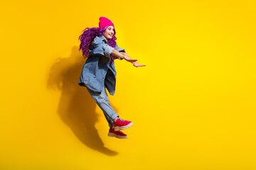 Young stylish woman with pink hair jumping joyfully in midair wearing casual denim clothes and a bright hat on a vibrant yellow background