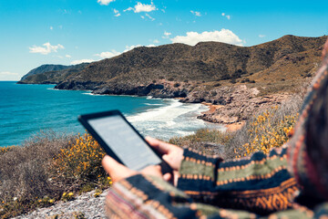 Close-Up of Person Holding eBook Reader Against Background of Rugged Calblanque Coastline and Hills in Murcia, Spain