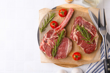 Pieces of raw beef, rosemary and tomatoes on white tiled table, flat lay. Space for text