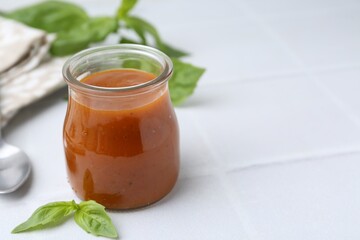 Tasty curry sauce in glass jar and basil on white tiled table, closeup. Space for text
