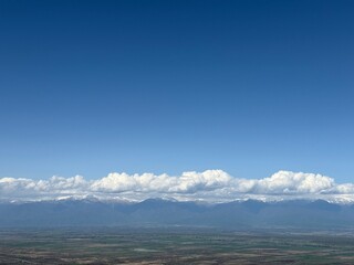 Snow-dusted mountains stand beneath a long row of fluffy clouds, with a vast, patchy green valley stretching out under a bright, deep blue sky