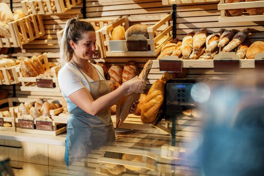 Saleswoman wearing an apron and plastic gloves is packing a loaf of bread into a paper bag, surrounded by various bread products displayed on wooden shelves in a bakery shop
