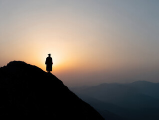 Silhouette of graduate standing on mountain peak at sunset