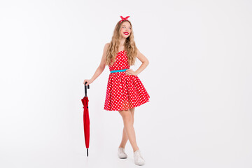 Charming young woman in a red polka dot dress posing with a retro umbrella on a white background showcasing vintage elegance.