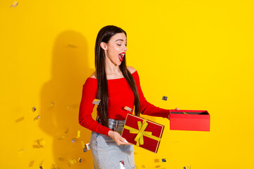 Smiling Young Woman in Red Shirt Holding an Open Gift Box Against a Vibrant Yellow Background with Confetti and a Cheerful Expression