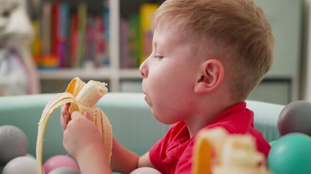 Close up of little boy in red shirt eating plantain while playing with colorful balls in pastel pit, hand holding almost finished banana visible in foreground, vibrant blurred background