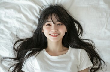 A young woman is lying on a bed, smiling warmly with a relaxed expression. The soft lighting highlights her long, dark hair and natural beauty, creating a serene and peaceful atmosphere.