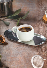 A detailed close-up of a ceramic white cup filled with black coffee, placed on a reflective mirror tray over a rustic stone-textured surface.