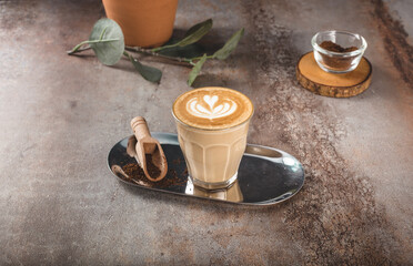 A close-up, eye-level shot of a glass of coffee latte with intricate heart-shaped latte art, placed on a reflective tray along with a wooden coffee scoop and coffee powder.