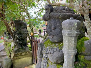 Ancient moss-covered stone statue with a serpent headdress in front of a traditional house in Java,...