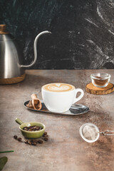 A white ceramic cup filled with latte featuring elegant heart tulip art, served on a reflective metal tray with a wooden scoop of ground coffee beside it.