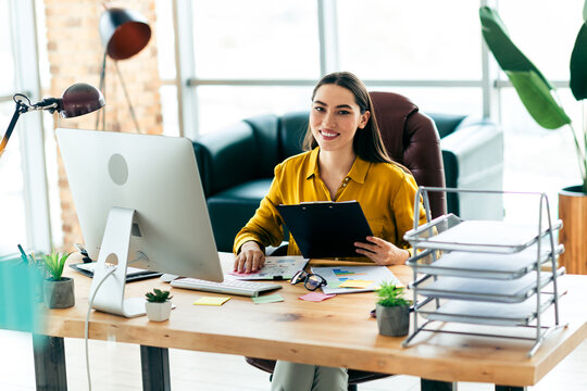 Confident businesswoman sitting at desk in bright modern office, holding clipboard for daily tasks and workplace management
