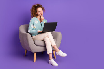 Fototapeta premium Young woman with red hair in a stylish cardigan using laptop on a chair against a vibrant purple background