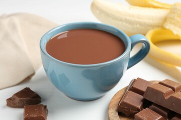 Tasty chocolate milk in cup, pieces and banana on white wooden table, closeup