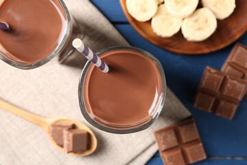 Tasty chocolate milk in glasses, pieces and banana on blue wooden table, flat lay