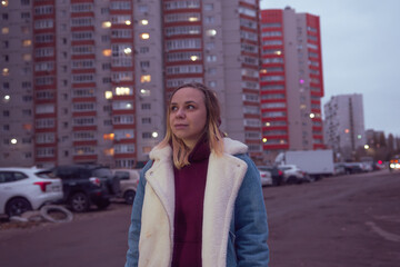 Fototapeta premium A young woman stands confidently in front of a modern residential building during dusk in an urban setting