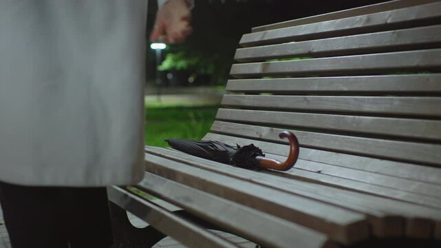 Back view of lady dropping umbrella on wooden bench then sitting calmly as she opens book to read under night park light, surrounded by greenery with soft streetlight bokeh in background