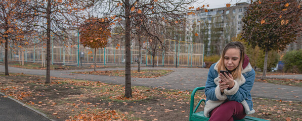 A woman dressed in a warm blue jacket sits on a bench in a city park, focused on her phone as...