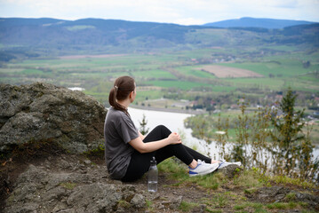 Woman in casual sportswear rests on top of hill, enjoying valley view. Great for wellness, motivation, solitude, endurance, personal growth and outdoor recovery.