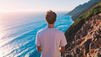 Fototapeta premium Back view of man in white t-shirt standing on cliff at sunset. Ocean view. Ideal for t-shirt mockup, branding, and lifestyle content. Inspirational and faceless concept.