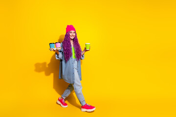 Young trendy woman with vibrant purple hair carrying colorful coffee cups on a bright yellow...
