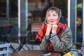 Fototapeta premium Woman enjoys a moment of reflection while seated outside a cafe on a chilly day with a warm scarf wrapped around her neck