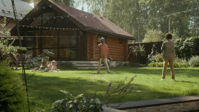 Wide shot of African American father playing baseball with teenage son outdoors on green lawn of suburban family home, boy throwing ball and dad catching, mother and sister relaxing on blanket
