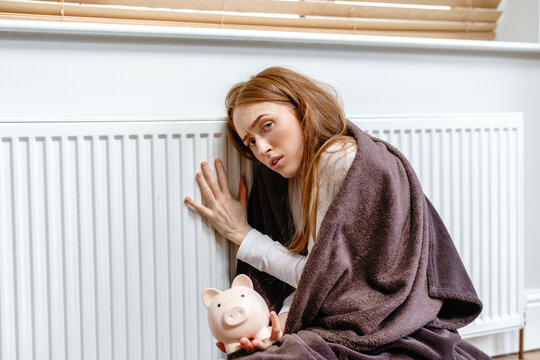 Young person sitting on  floor while wrapped in  blanket, holding  piggy bank, facing  radiator in cozy indoor setting during chilly weather
