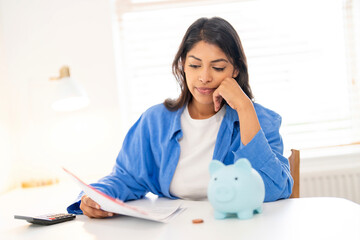 Woman analyzing financial documents while contemplating savings with a piggy bank on a desk in a...